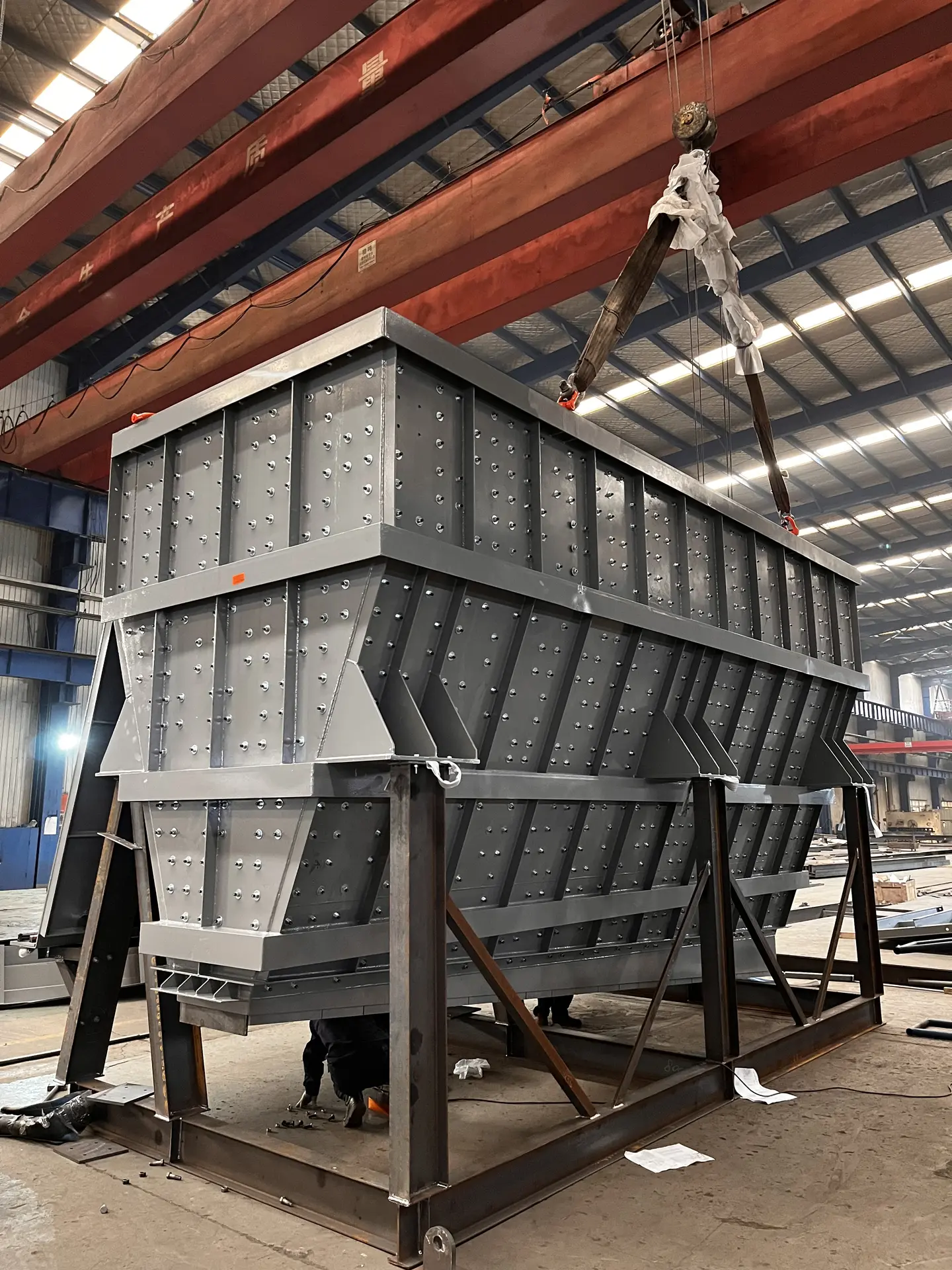 Grey steel hopper suspended from a crane hook inside the workshop